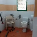 Baño adaptado con inodoro elevado, asideros laterales y silla de ducha